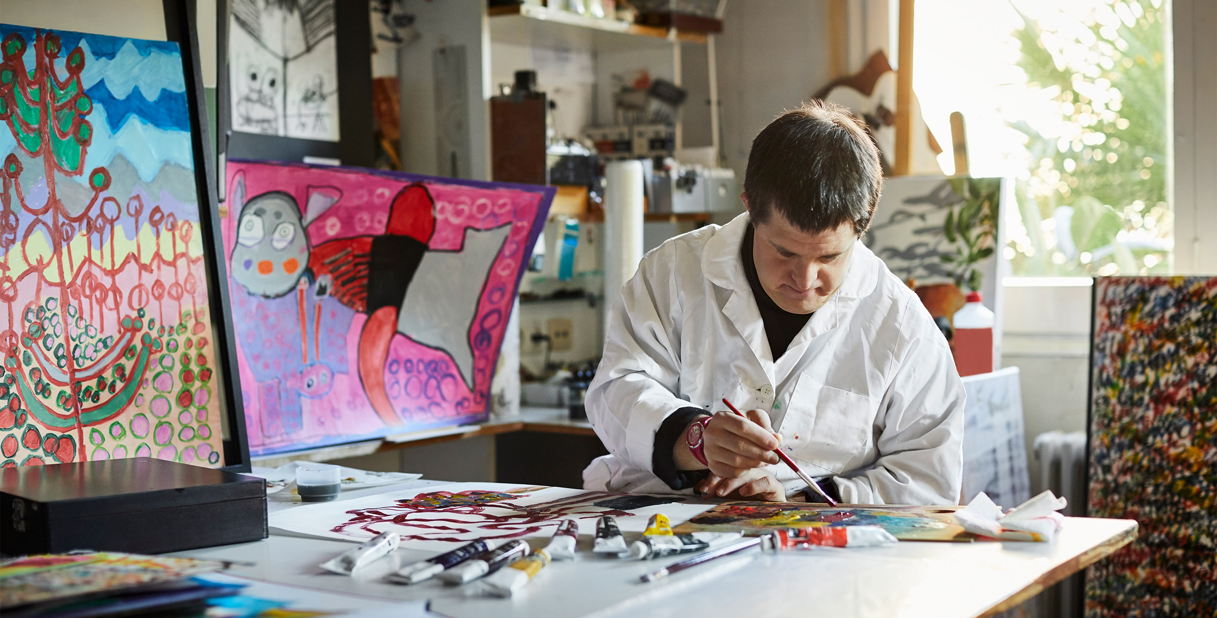 Das Foto zeigt eine Person in einem weissen Kittel, die gerade mit Pinsel und Farben malt.
